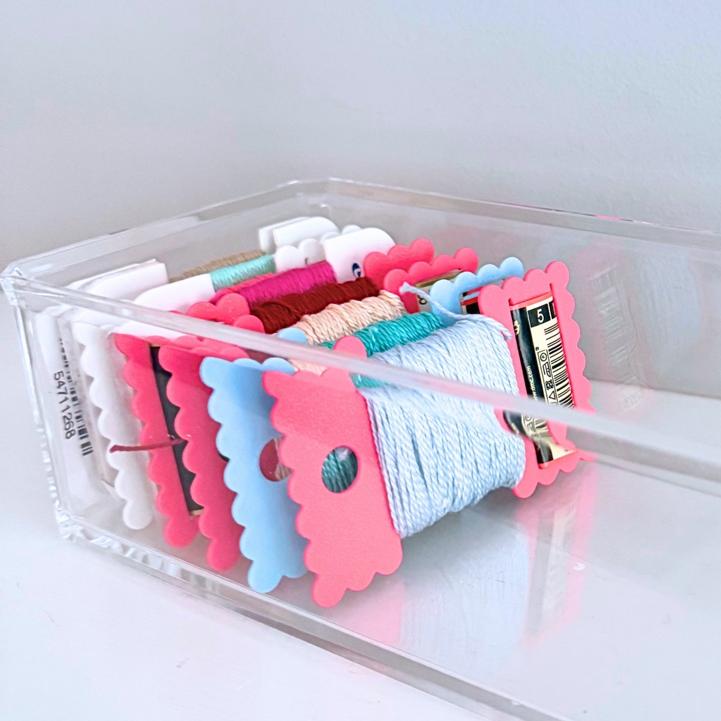 A clear acrylic organizer tray holding multiple colorful skeins of thread in pink, blue, white, teal, and coral, photographed on a clean white surface against a light gray background.