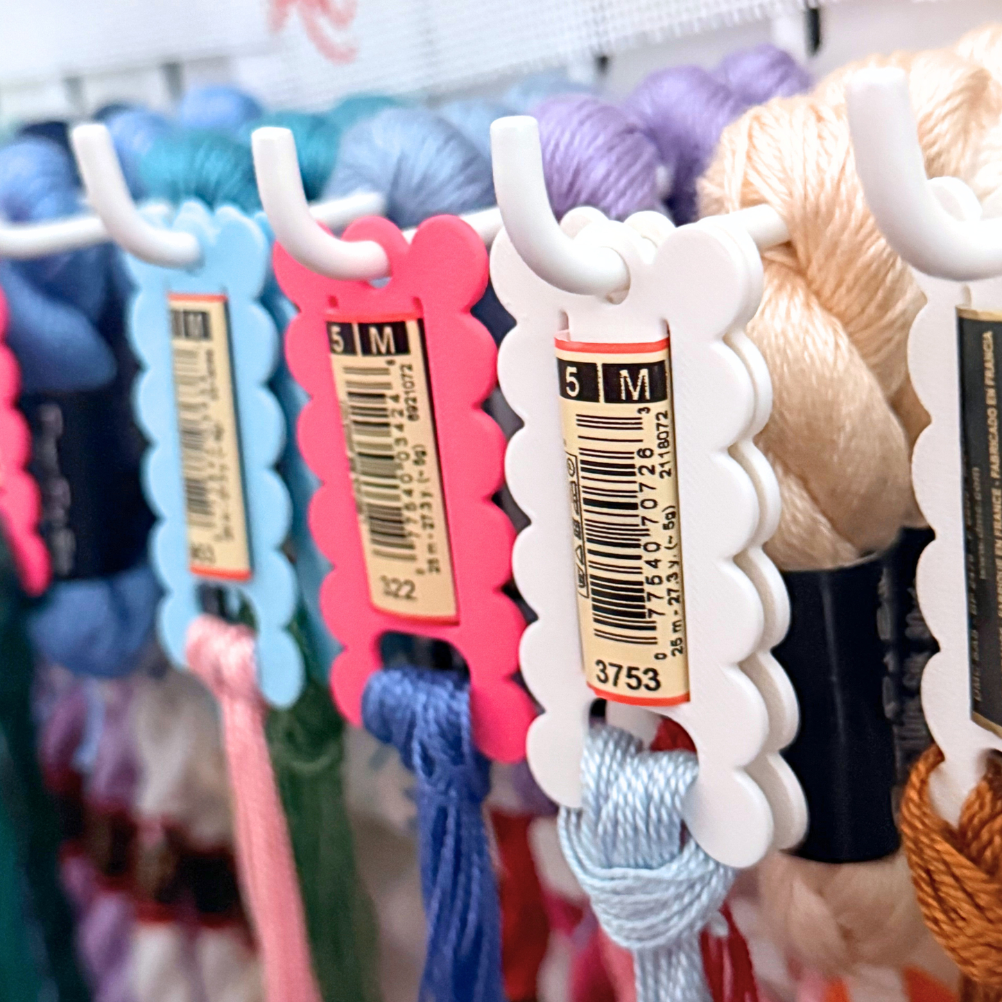 Close-up of a display of colorful needlepoint skeins hanging on white hooks, with product tags attached. The skein card comes in a variety of colors including blue, pink, purple, beige, and orange.