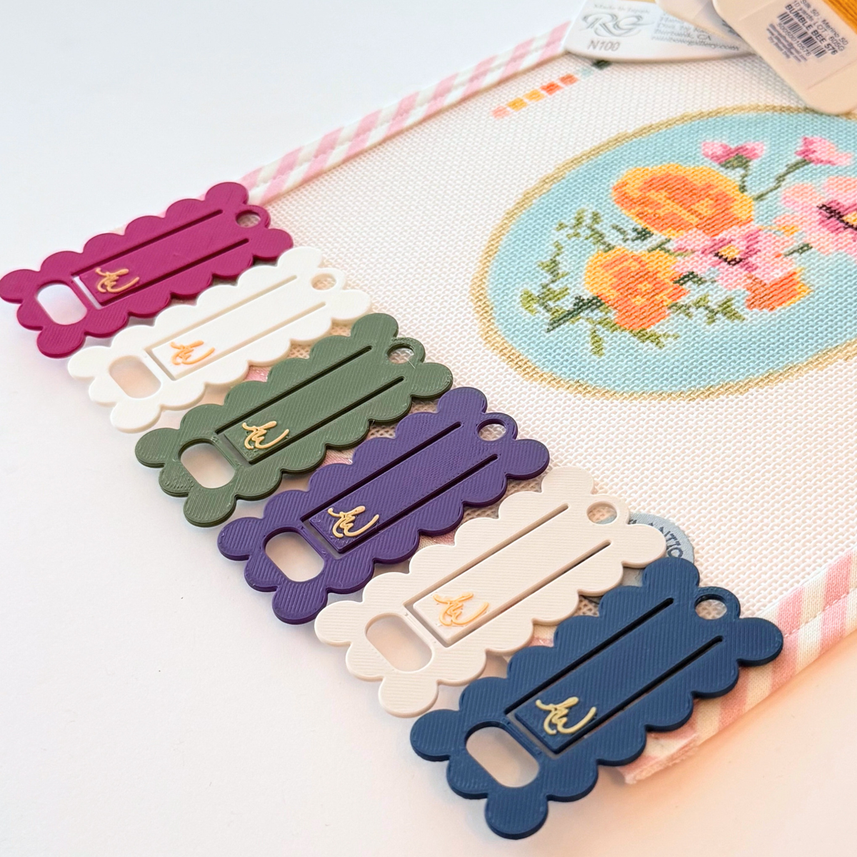 A close-up of needlepoint floss ThreadKeeper™️ floss drops displayed on a white needlepoint canvas with pink stripe trim. The floss includes green, navy, white, grey, plum and indigo, on a gold ring.