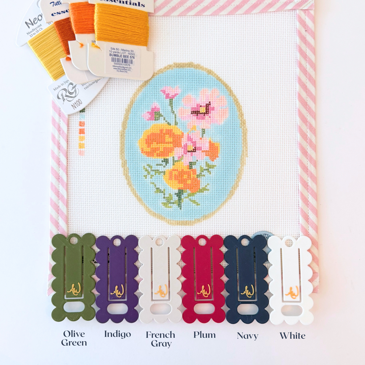 A close-up of needlepoint floss ThreadKeeper™️ floss drops labeled with their colors on a white needlepoint canvas with pink stripe trim. The floss includes green, navy, white, grey, plum and indigo.