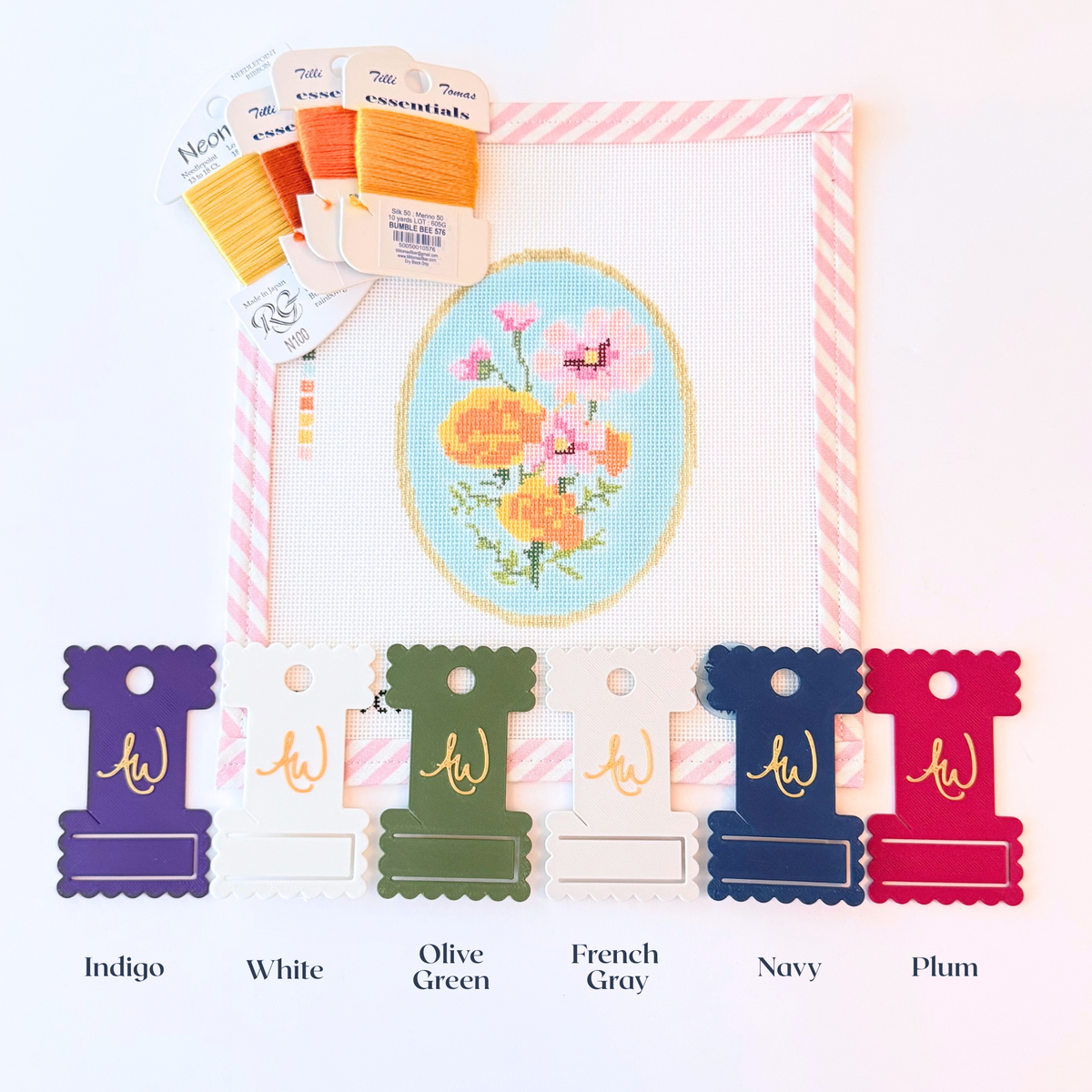 A close-up of needlepoint ThreadKeeper™️ thread cards displayed on a white needlepoint canvas with pink stripe trim. The floss includes are labeled with their color names olive green, navy, white, grey, plum and indigo, on a gold ring.