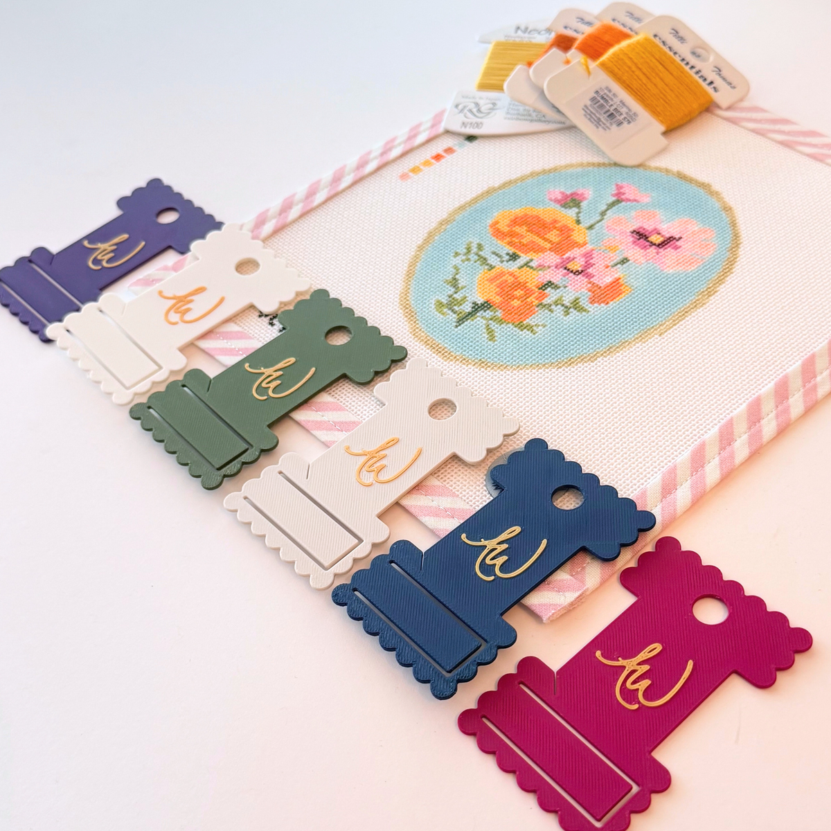 A close-up of needlepoint ThreadKeeper™️ thread cards displayed on a white needlepoint canvas with pink stripe trim. The floss includes green, navy, white, grey, plum and indigo.