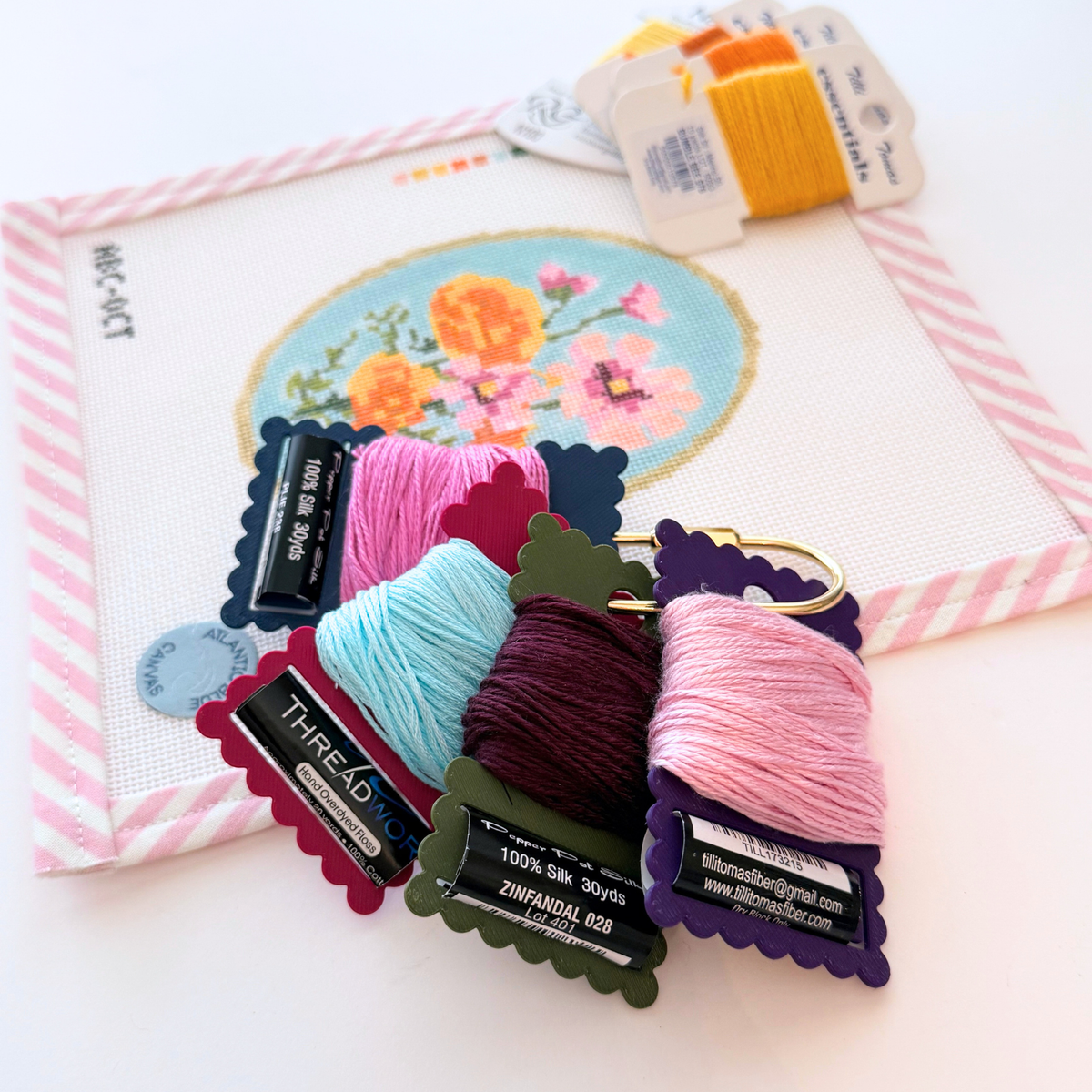 A close-up of needlepoint ThreadKeeper™️ thread cards displayed on a white needlepoint canvas with pink stripe trim. The floss includes green, navy, white, grey, plum and indigo, on a gold ring.