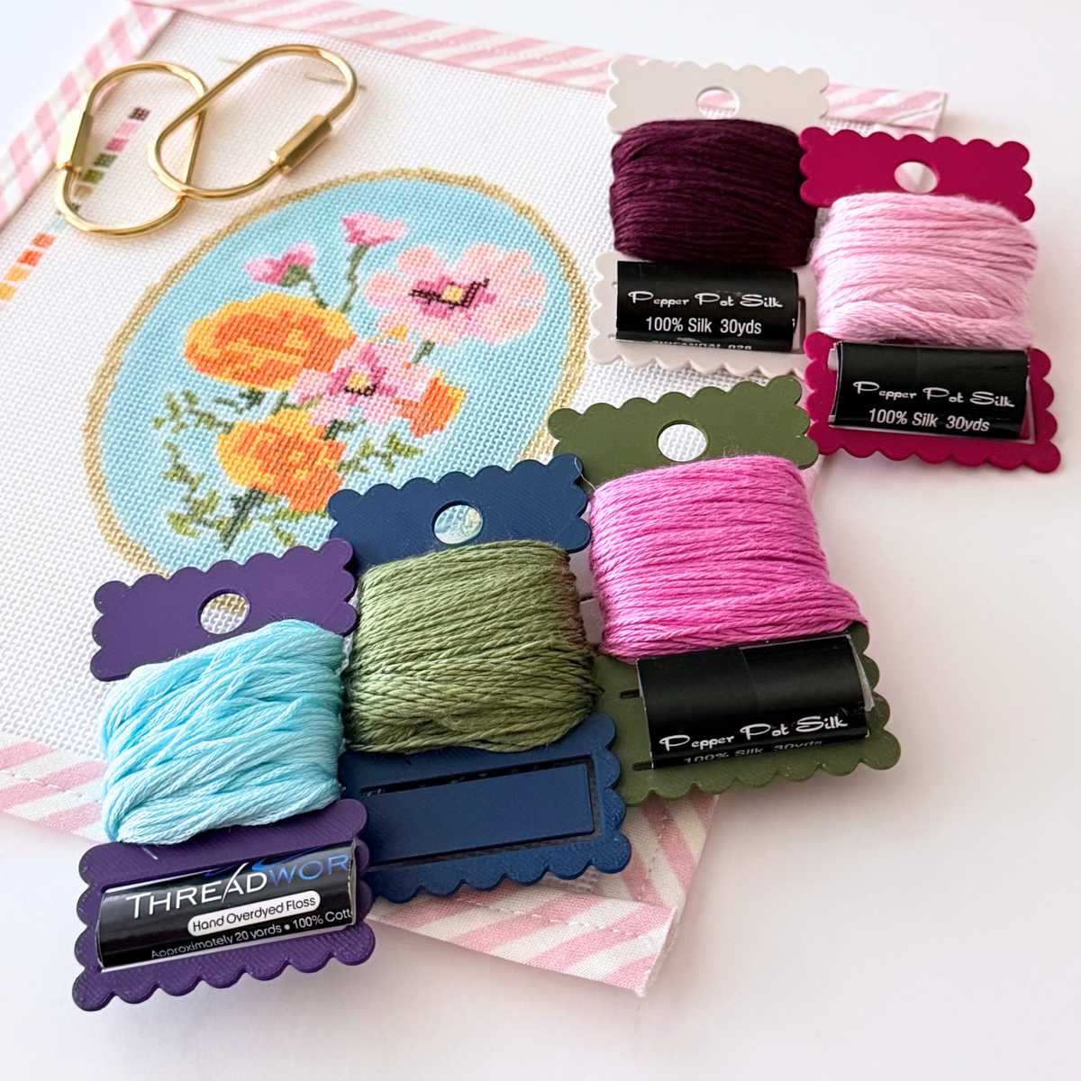 A close-up of needlepoint ThreadKeeper™️ thread cards displayed on a white needlepoint canvas with pink stripe trim. The floss includes green, navy, white, grey, plum and indigo, on a gold ring.