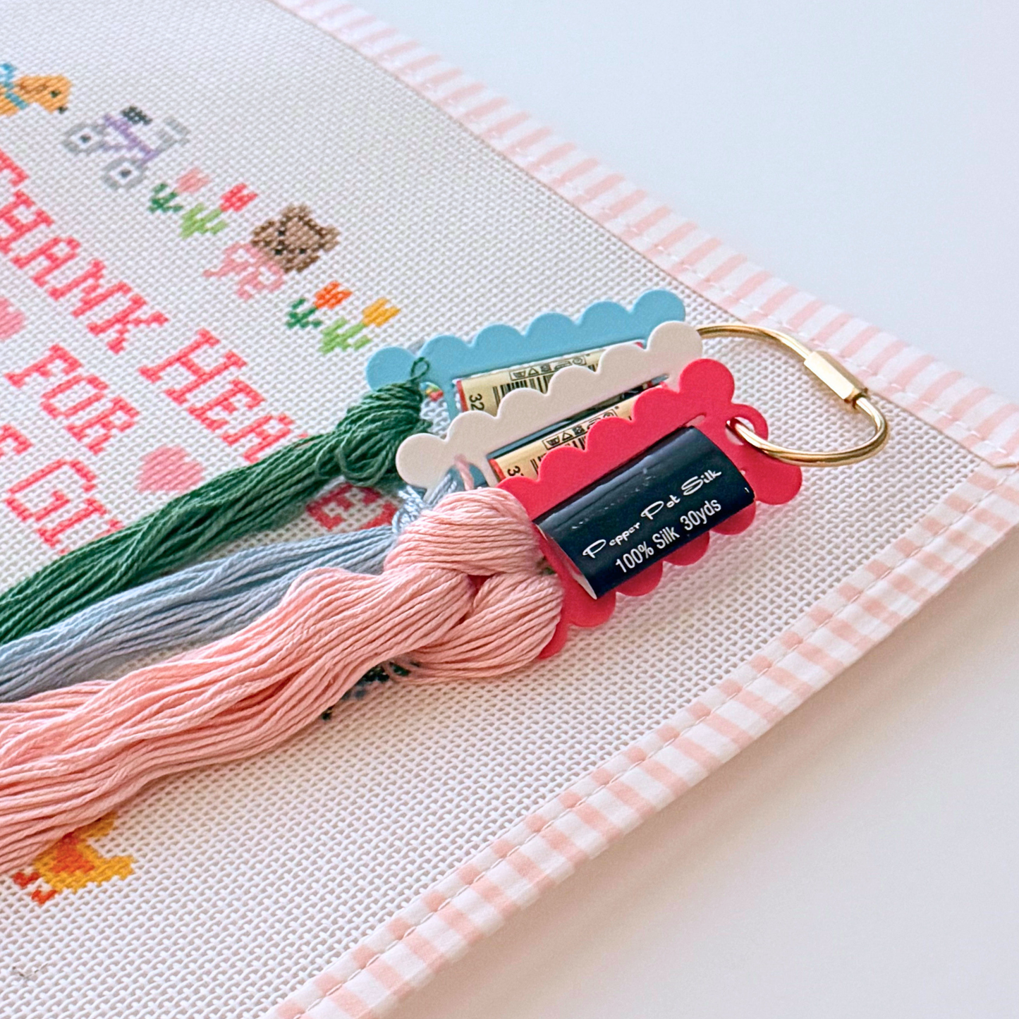 A close-up of a handmade needlepoint sampler with a pink and white gingham border, featuring a partially finished needlepoint design.