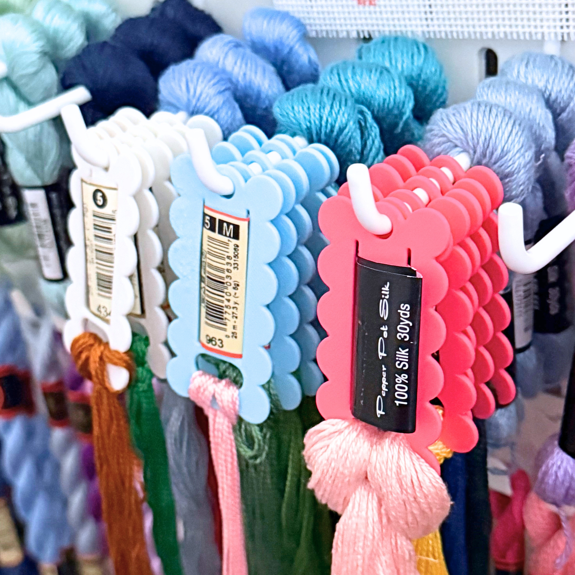 A close-up of hanging skeins of thread in multiple colors, with scalloped floss drop tags showing skein details like color and length. 