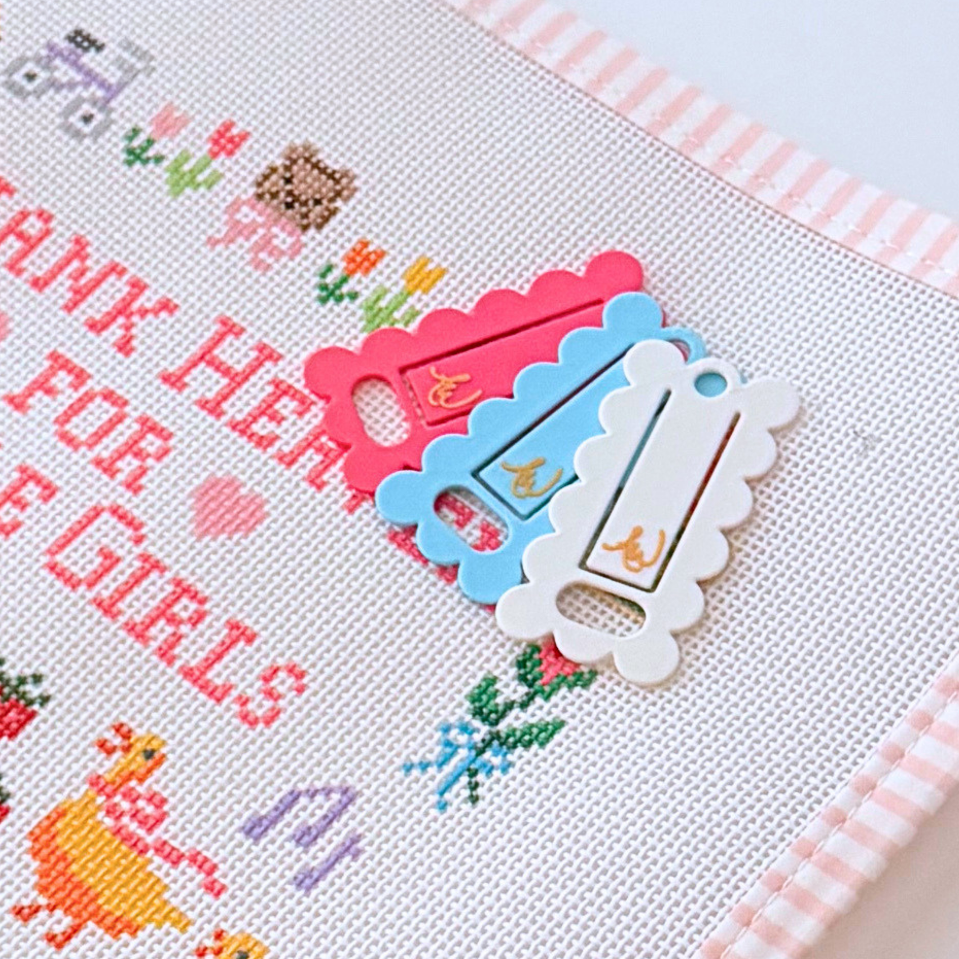 Three scalloped needlepoint floss drops in pink, light blue, and white. Floss drops are shown in pink, blue and white and arranged on a needlepoint canvas.