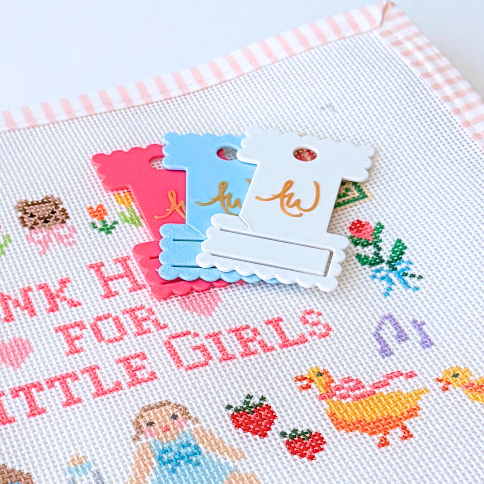 A close-up of a white needlepoint canvas with embroidered text reading 'THANK HEAVEN FOR LITTLE GIRLS' and small colorful stitched motifs, with three decorative scallop thread cards placed on top.