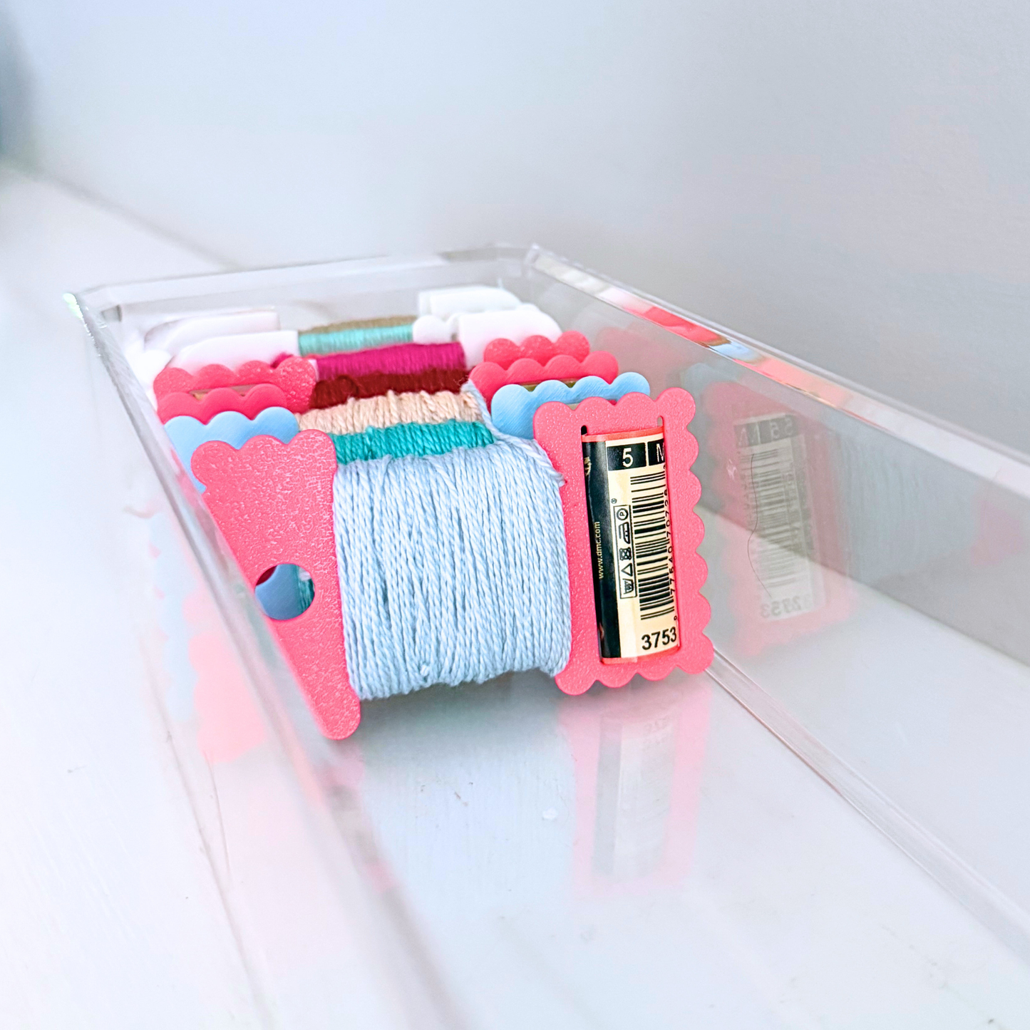 A clear plastic tray holding multiple skeins of thread in assorted colors, with a light blue spool in front and pink scalloped ends on the spools.