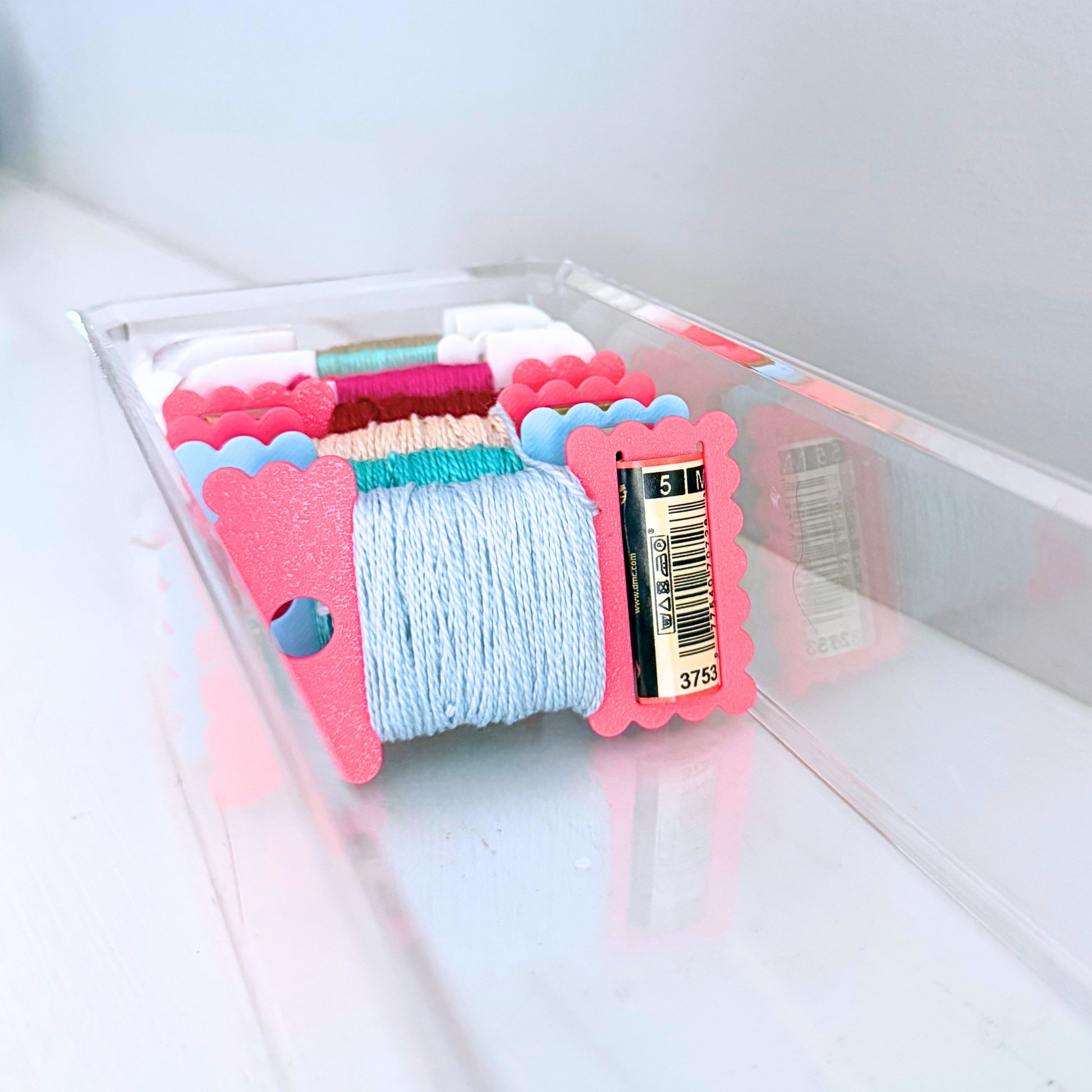 A clear plastic tray holding multiple skeins of thread in assorted colors, with a light blue spool in front and pink scalloped ends on the spools.