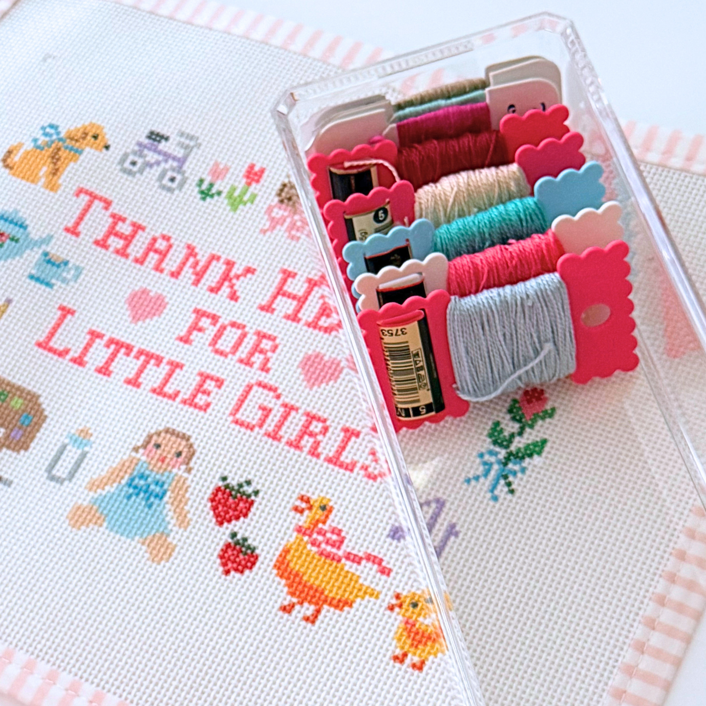 A close-up of a needlepoint canvas with the words “THANK HEAVEN FOR LITTLE GIRLS” stitched in pink, alongside a clear plastic case holding multiple skeins of colorful thread.