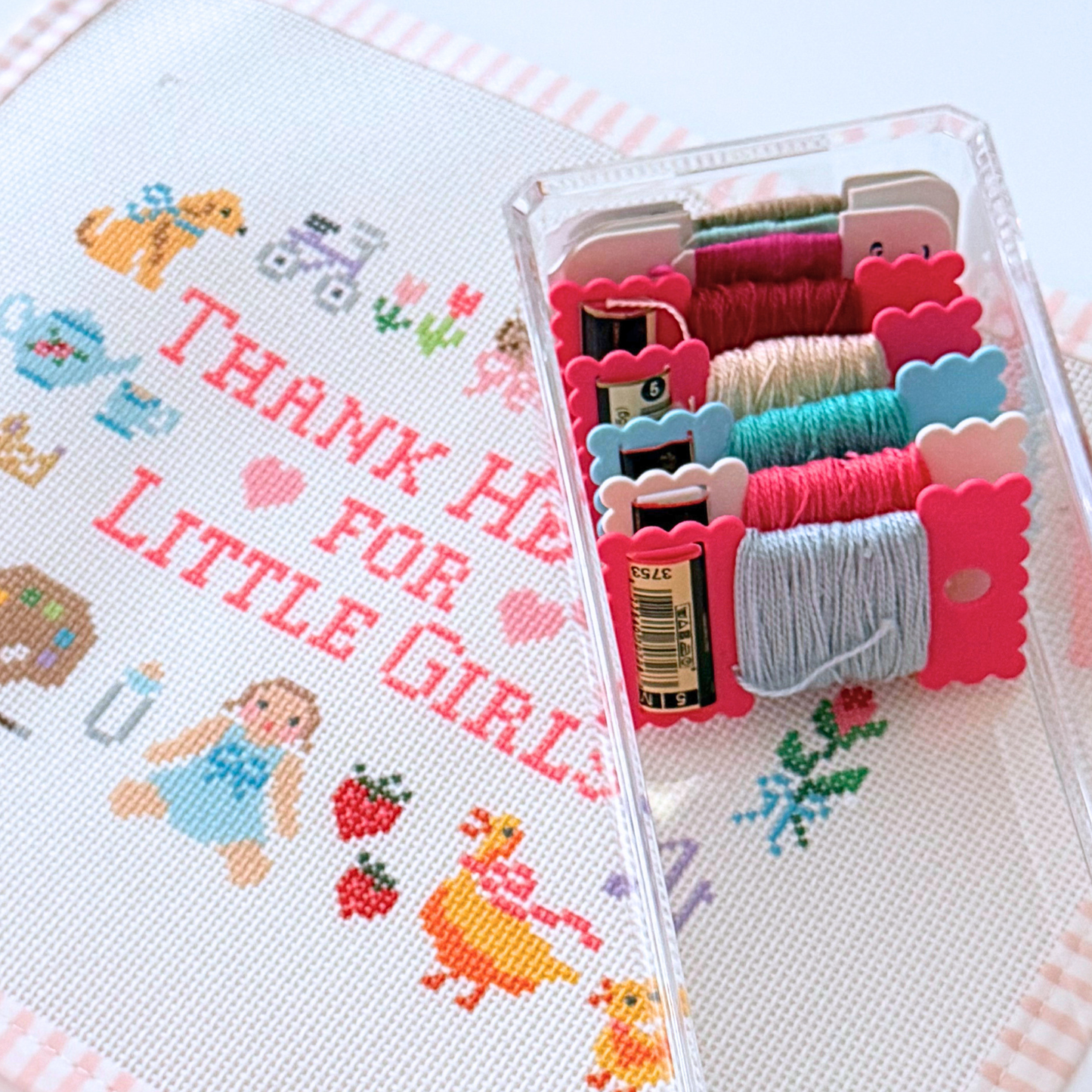 A close-up of a needlepoint kit showing a canvas with the text 'THANK HEAVEN FOR LITTLE GIRLS' and whimsical characters, alongside a clear plastic case holding multiple skeins of colorful thread.