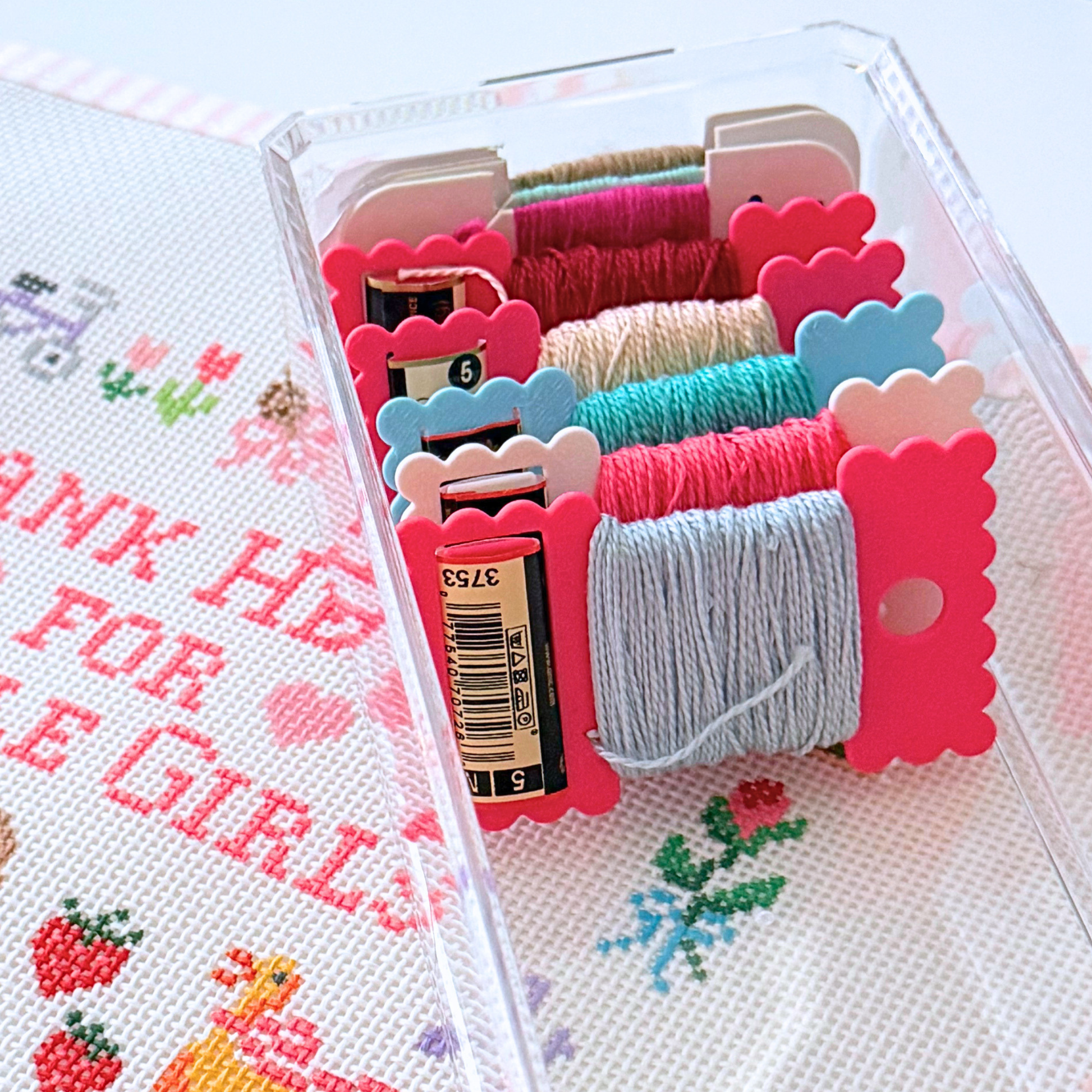 A clear plastic organizer case holding multiple spools of needlepoint skeins in bright pink, blue, teal, beige, and gray, placed on top of a white needlepoint canvas with pink lettering and colorful floral motifs.
