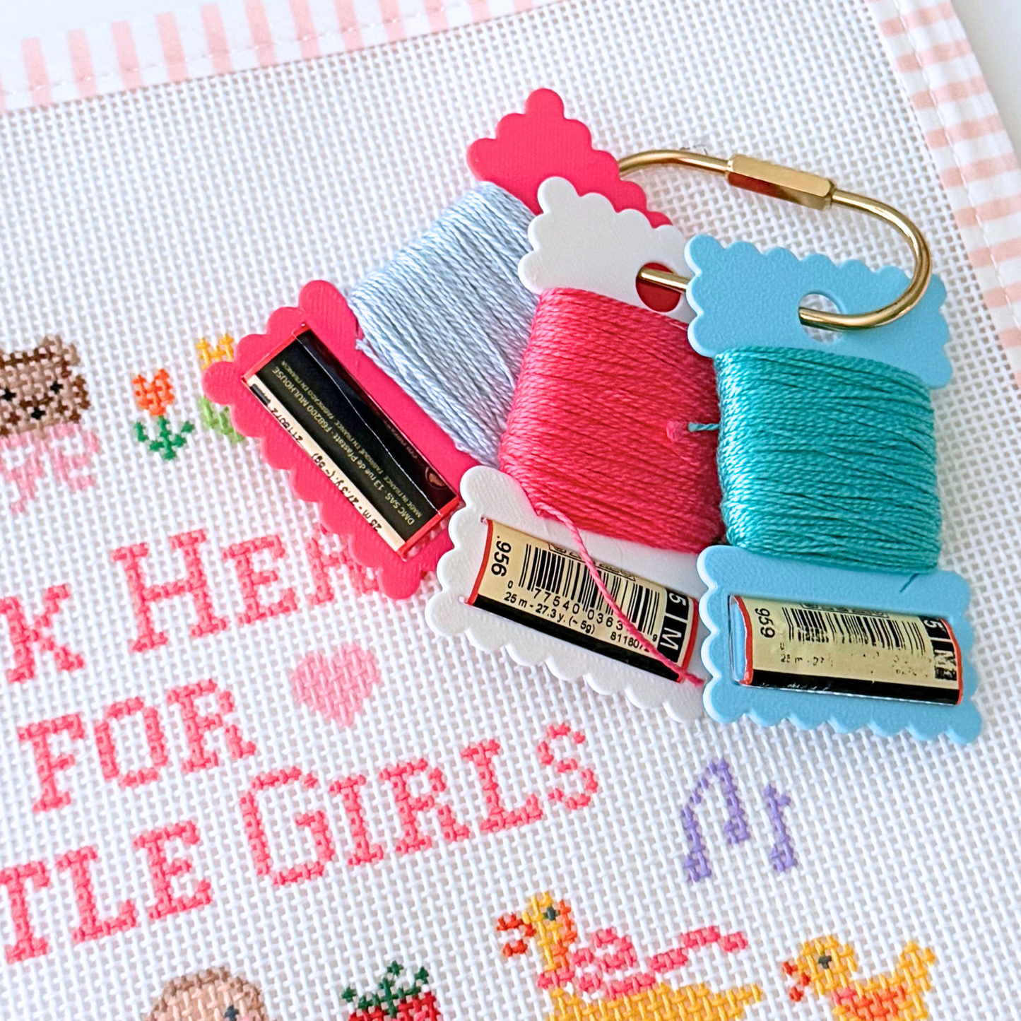 Three skeins of needlepoint thread in light blue, pink, and teal, attached to a gold-ring organizer, displayed on a white needlepoint canvas with pink lettering.