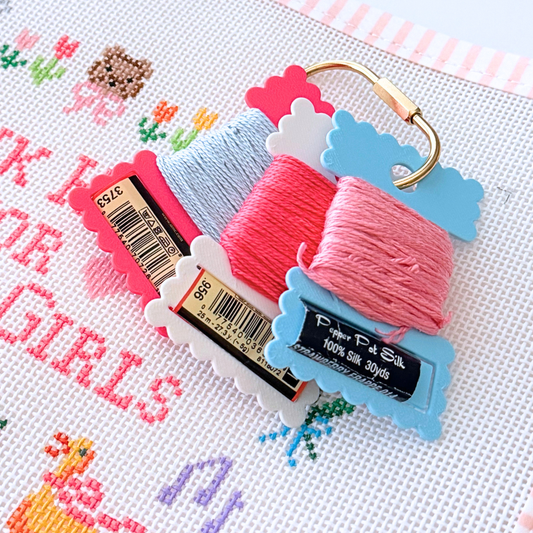 Three skeins of needlepoint thread in light blue, coral pink, and medium pink, attached to scalloped paper hangers with a gold ring clasp, displayed on white needlepoint canvas with a design.