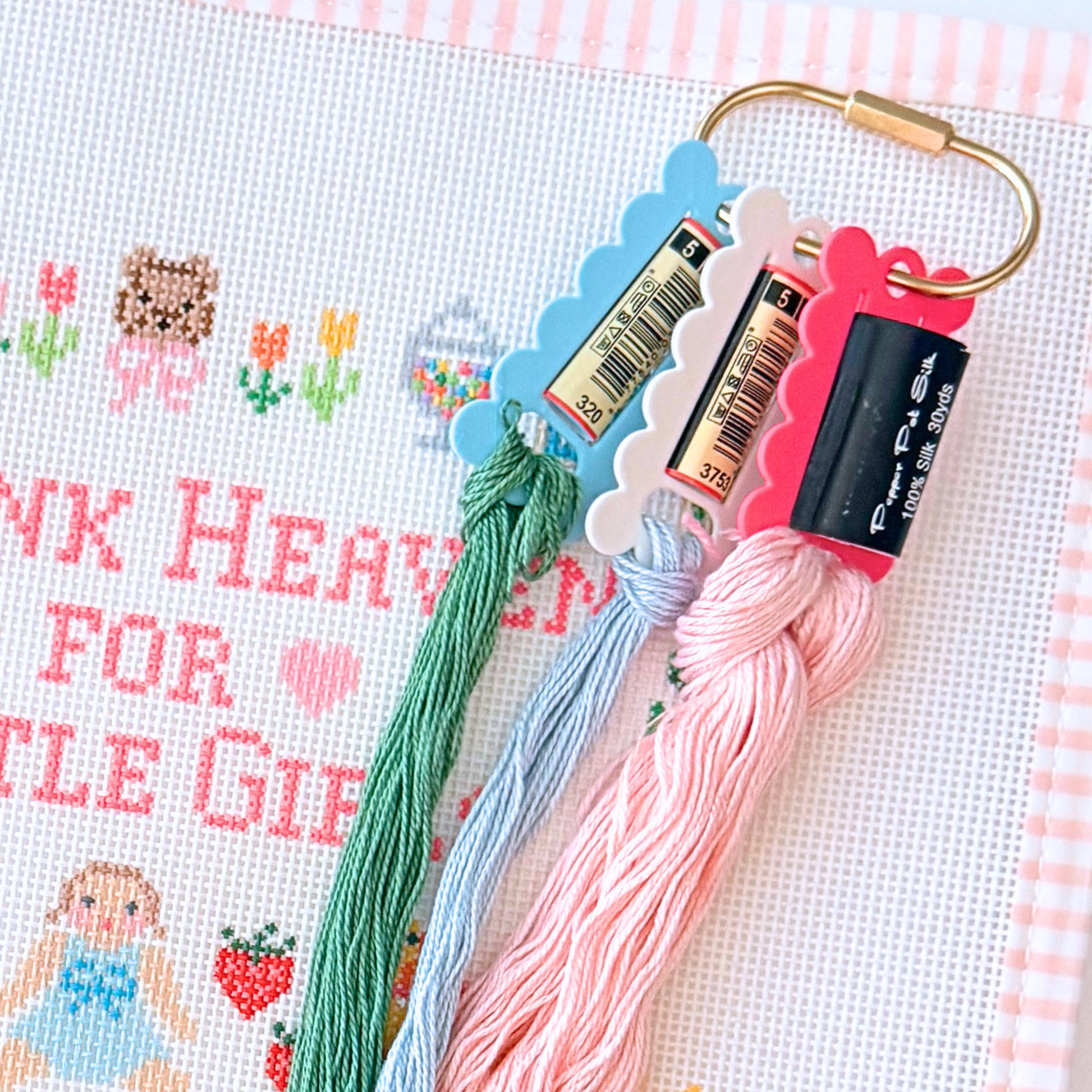 A close-up of needlepoint floss skeins attached to a gold ring, displayed on a white needlepoint canvas with pink gingham trim. The floss includes green, light blue, and light pink colors, on a gold ring.